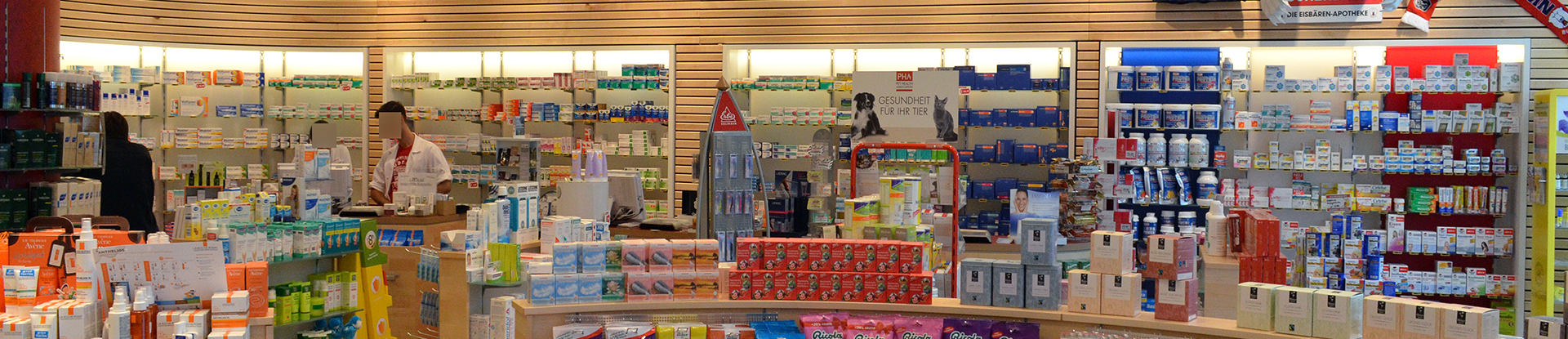 Innenansicht einer Apotheke. Es sind mehrere Regale an der Wand sowie im Verkaufsraum mit Medikamenten. Im Hintergrund stehen zwei Mitarbeiter mit unkenntlichen Gesichtern an der Kasse.