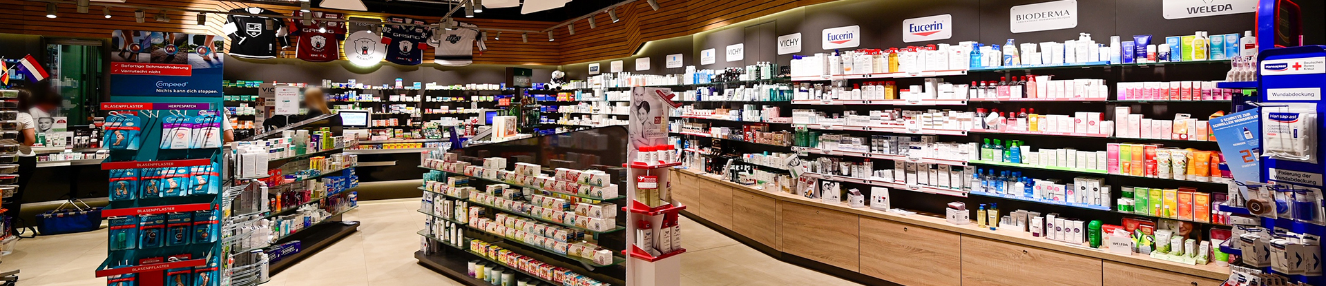 Innenansicht der Arena Apotheke. Regale an der Wand & im Verkaufsraum mit Medikamenten. An der Wand hängen Eishockey-Trikots