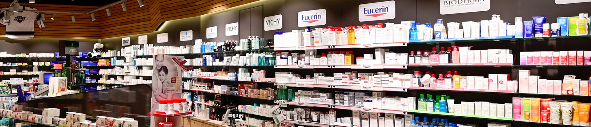 Innenansicht der Arena Apotheke. Hohe Regale an der Wand mit Kosmetikprodukten verschiedener Marken, deren Logos darüber hängen.