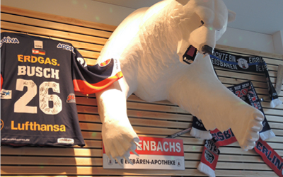 Eine Statue eines Eisbären-Oberkörper hängt and einer vertäfelten Wand. Darum herum hängen Eishockey-Schals und ein Schild mit Aschenbachs, die Eisbären Apotheke
